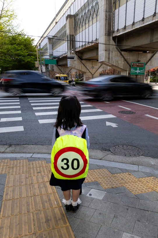 어린이가 '제한속도 30km'임을 보여주는 가방을 메고 신호를 기다리고 있다. (사진 출처: 시사IN)