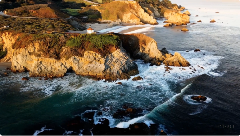 Prompt: Drone view of waves crashing against the rugged cliffs along Big Sur’s garay point beach. The crashing blue waters create white-tipped waves, while the golden light of the setting sun illuminates the rocky shore. A small island with a lighthouse sits in the distance, and green shrubbery covers the cliff’s edge. The steep drop from the road down to the beach is a dramatic feat, with the cliff’s edges jutting out over the sea. This is a view that captures the raw beauty of the coast and the rugged landscape of the Pacific Coast Highway.